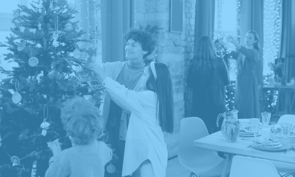 Group of people (family) gathered together for Christmas. One lady is decorating a tree with a little girl and boy. In the background two other women are speaking to eachother.